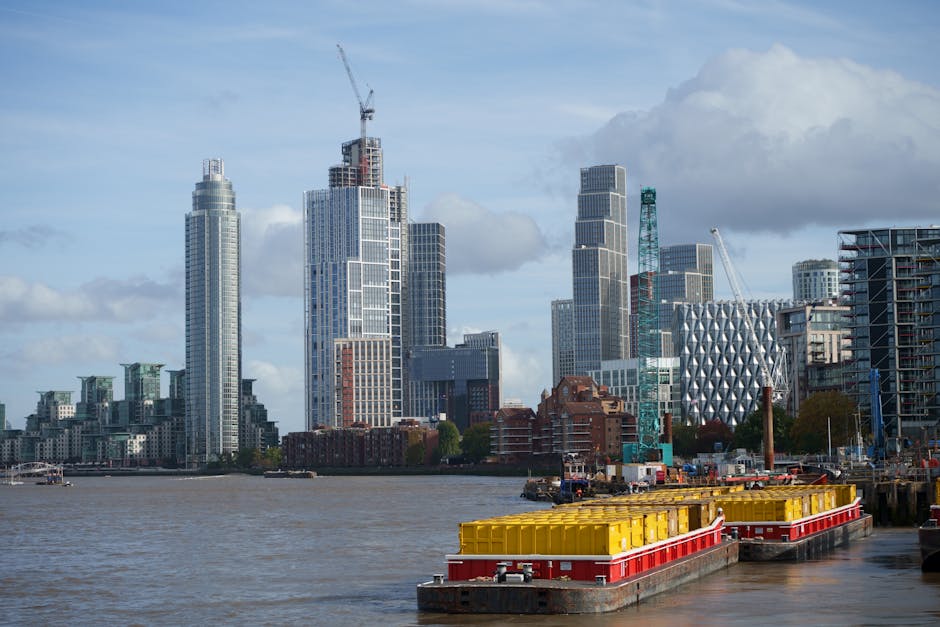A modern red London Underground train traveling across a bridge over the River Thames in Docklands, surrounded by tall contemporary office buildings with glass facades. The scene is set during dayligh