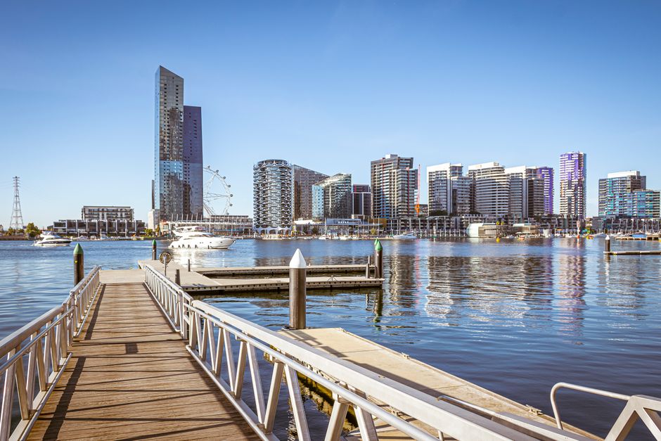 A waterfront scene in Docklands with a wooden dock extending over the water, enclosed by temporary fencing and safety barriers. Several people are visible, some inspecting the area and others standing
