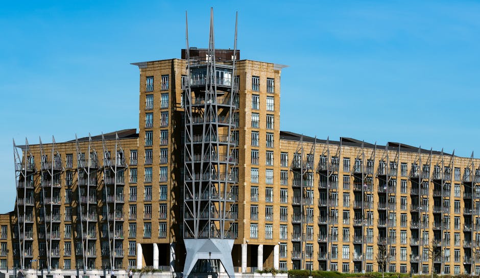 A large multi-story residential building with a modern architectural design features numerous balconies with metal railing details. The structure is constructed with light brown bricks and has a prominent central tower with a steel framework extending above the roof, possibly for communication or maintenance purposes. In the foreground, a paved area and sidewalk are visible, with no furniture or interior elements seen. The clear blue sky provides natural lighting that highlights the building's exterior textures and shadows cast by the protruding elements. This setting suggests the building is located in the Docklands area, fitting a context of house removals, moving logistics, or furniture transport activities associated with relocations. The image is relevant to professional removals services offered by Movers Docklands, emphasizing the urban environment where relocating furniture or household items may involve navigating such large structures during the loading process.