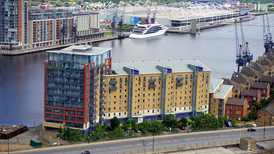 A large multi-story residential building with a modern architectural design features numerous balconies with metal railing details. The structure is constructed with light brown bricks and has a prominent central tower with a steel framework extending above the roof, possibly for communication or maintenance purposes. In the foreground, a paved area and sidewalk are visible, with no furniture or interior elements seen. The clear blue sky provides natural lighting that highlights the building's exterior textures and shadows cast by the protruding elements. This setting suggests the building is located in the Docklands area, fitting a context of house removals, moving logistics, or furniture transport activities associated with relocations. The image is relevant to professional removals services offered by Movers Docklands, emphasizing the urban environment where relocating furniture or household items may involve navigating such large structures during the loading process.