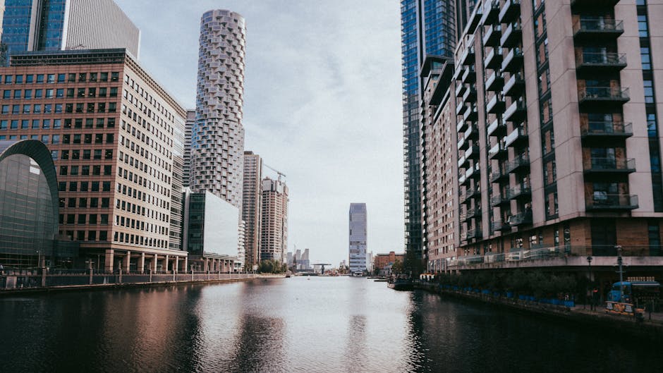 A wide view of the Docklands area in Canary Wharf featuring tall modern skyscrapers lining the sides of a wide river or canal. The buildings exhibit various architectural styles, including glass facades, steel structures, and brick exteriors, with some under construction and crane machinery visible. The water reflects the buildings and the partly cloudy sky above. On the right, there are residential buildings with balconies, while on the left, commercial office towers dominate the skyline. The scene captures the urban environment typical of a busy city centre, suitable for house removals and furniture transport activities by Movers Docklands. No people, vehicles, or moving equipment are visible in this shot, emphasizing the architectural landscape of this commercial and residential district, often involved in home relocation or moving services.