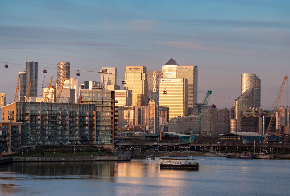 A panoramic view of the Canary Wharf financial district skyline in Docklands during the late afternoon or early evening, with the sunlight casting a warm glow on the modern glass and steel high-rise office buildings, including structures branded with Barclays. Several construction cranes are visible among the buildings, indicating ongoing development, and a cable car system with red gondolas is suspended above the river in the foreground. At the base of the image, a river reflects the cityscape, with some boats docked along the quay. To the left, a residential building featuring balconies is positioned near the water, while outside, a loading or unloading area might be used for moving furniture and household items typical in home relocation projects. This setting exemplifies an urban environment where professional removals services, like those offered by Movers Docklands, facilitate the logistics of furniture transport and packing during house moves in the Canary Wharf E14 area.