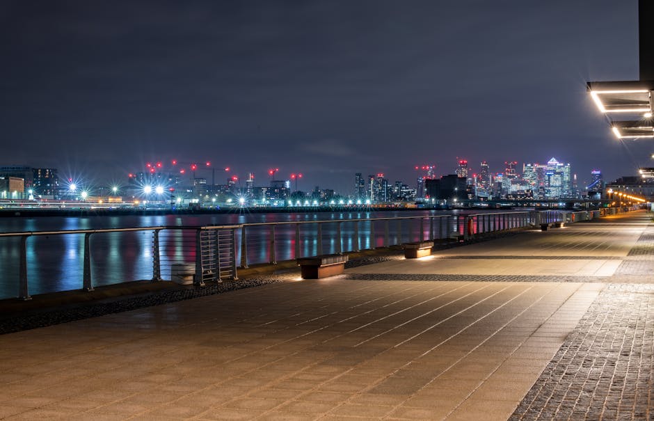 A wide view of the Docklands area in Canary Wharf featuring tall modern skyscrapers lining the sides of a wide river or canal. The buildings exhibit various architectural styles, including glass facades, steel structures, and brick exteriors, with some under construction and crane machinery visible. The water reflects the buildings and the partly cloudy sky above. On the right, there are residential buildings with balconies, while on the left, commercial office towers dominate the skyline. The scene captures the urban environment typical of a busy city centre, suitable for house removals and furniture transport activities by Movers Docklands. No people, vehicles, or moving equipment are visible in this shot, emphasizing the architectural landscape of this commercial and residential district, often involved in home relocation or moving services.